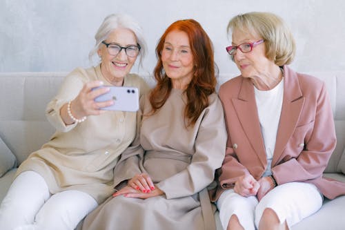 Free Senior cheerful women using mobile phone and looking at screen while making video call together while resting on sofa together Stock Photo