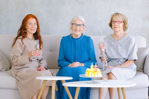 Aged cheerful women smiling and celebrating birthday while looking at camera at table with sweet cake decorated with candles
