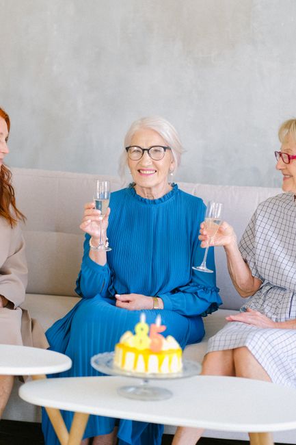 Group of elderly cheerful women with glasses of champagne celebrating birthday at table with tasty sweet cake