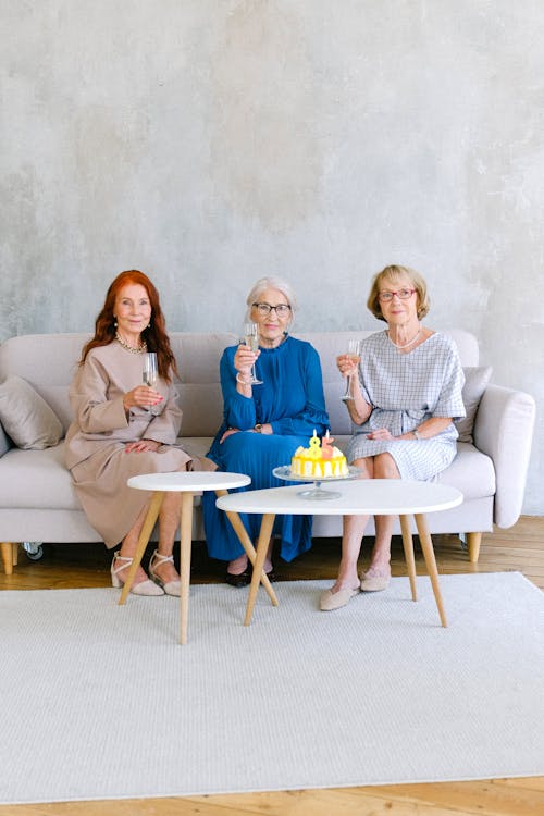 Full body of cheerful aged women with glasses of champagne at table with birthday cake decorated with candles