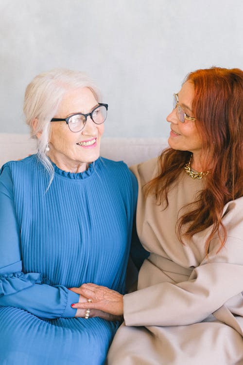 Senior cheerful women in eyeglasses and trendy dresses holding hands while smiling and talking