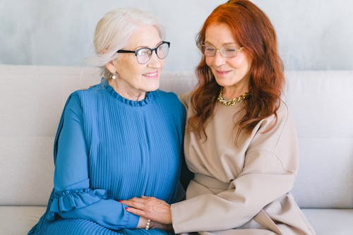 Positive elderly women in eyeglasses and elegant trendy dressing holding hands and speaking while resting on sofa