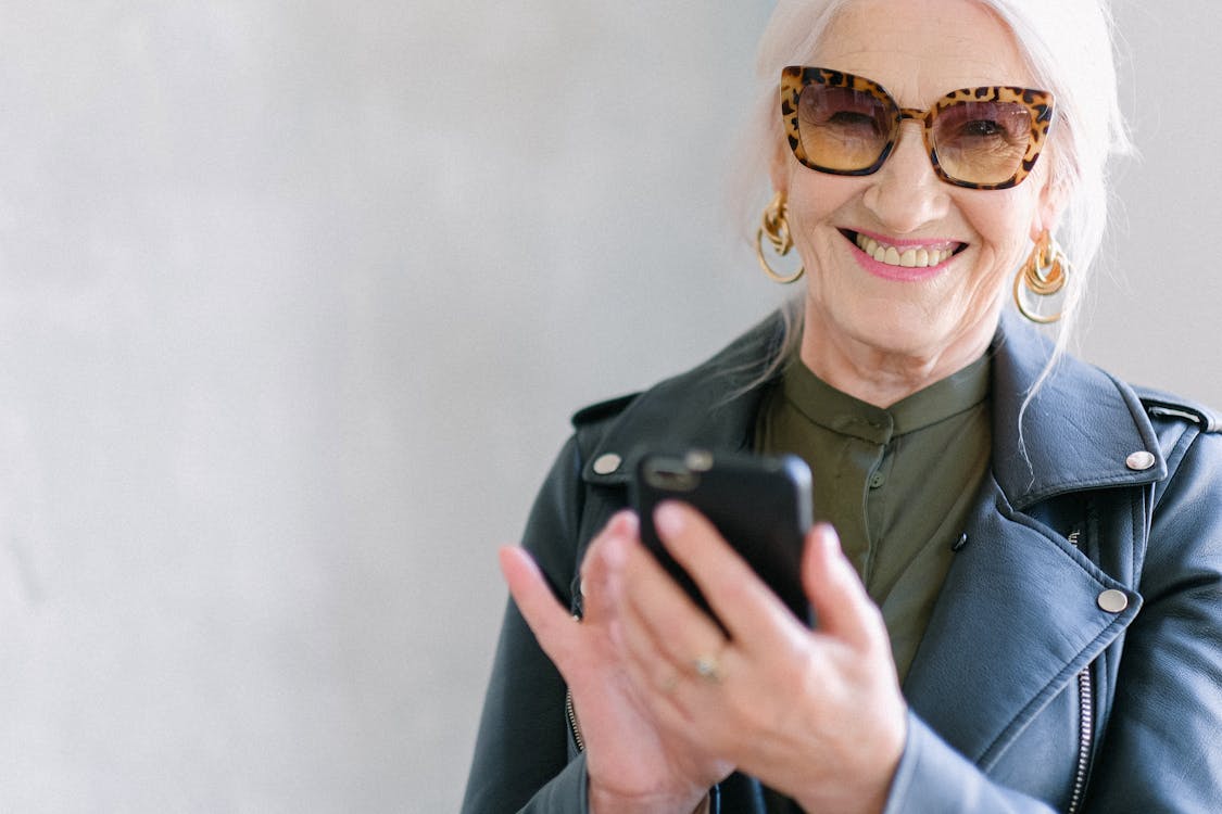 Crop happy senior female in stylish jacket touching screen of mobile phone while smiling and looking at camera