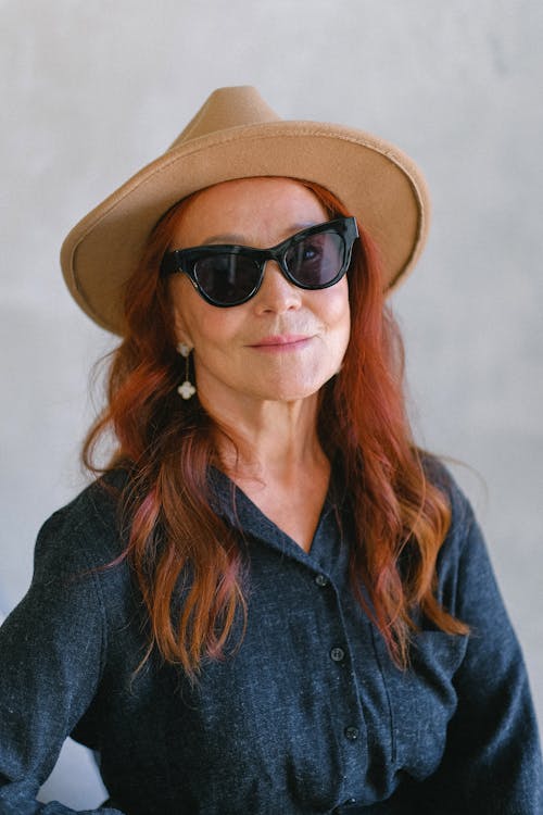 Elderly stylish female in fashionable sunglasses and hat looking at camera while smiling on gray background