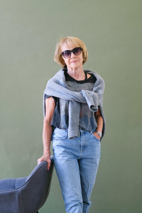 Aged female in sunglasses and stylish outfit leaning on armchair and looking at camera on green background of studio