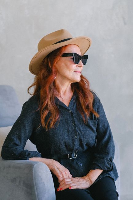 Elegant woman with red hair wearing a hat and sunglasses, exuding a chic fashion style against a neutral background.