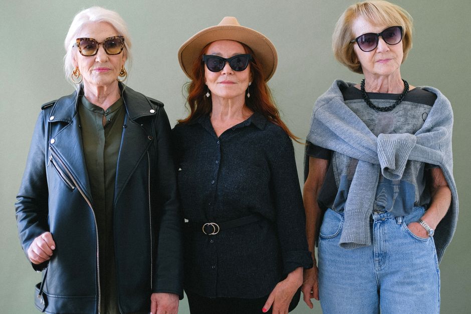 Aged fashionable women in sunglasses and stylish outfit looking at camera on green background