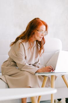 Aged concentrated female with red hair in eyeglasses surfing internet on netbook in modern room