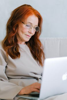 A senior woman with red hair and eyeglasses types on a laptop indoors.