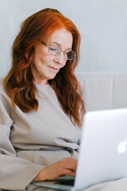 Free A senior woman with red hair and eyeglasses types on a laptop indoors. Stock Photo