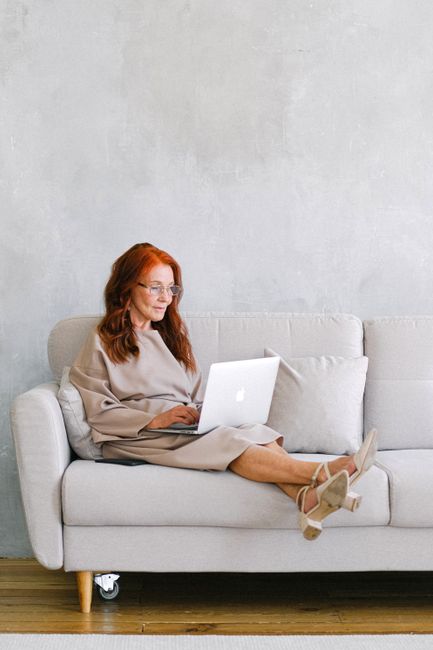 A senior woman with glasses sits on a sofa using a laptop for remote work, embodying the digital nomad lifestyle.