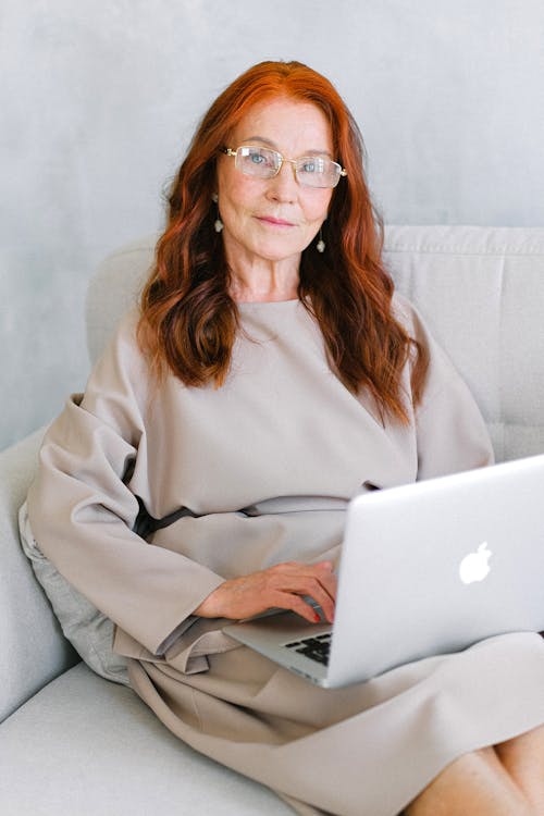 Free Cheerful senior woman entrepreneur with long red hair in dress sitting on comfortable sofa and browsing laptop at home Stock Photo