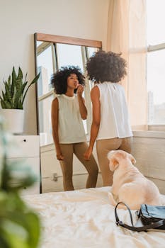 Young woman applies makeup in a bright room with her dog watching from the bed.