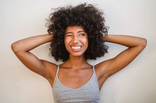 Free Expressive African American female with Afro hairstyle holding hands behind head while standing near wall in studio and looking away Stock Photo