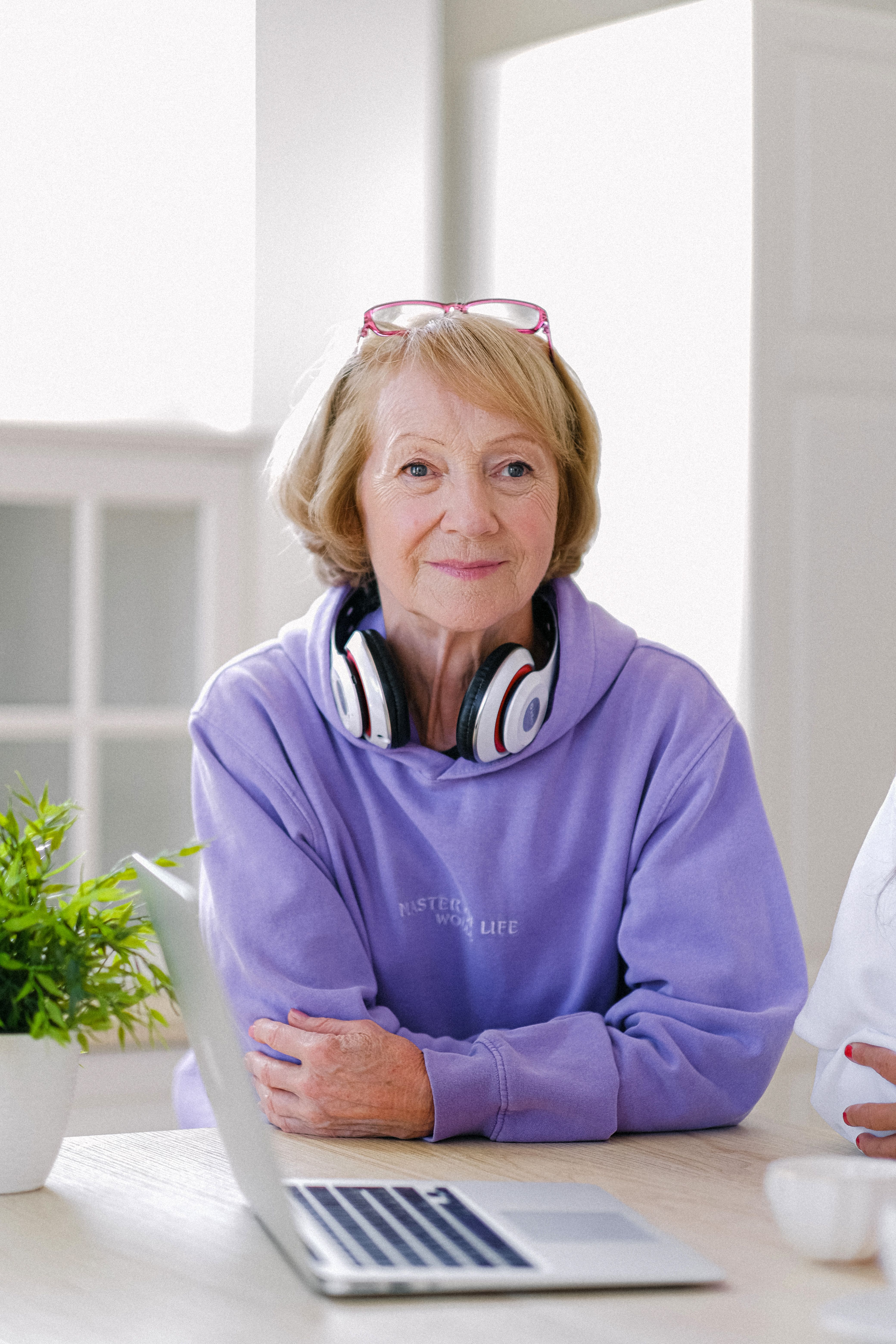 Cheerful senior woman with headphones in stylish outfit sitting at table with netbook in light apartment in daytime