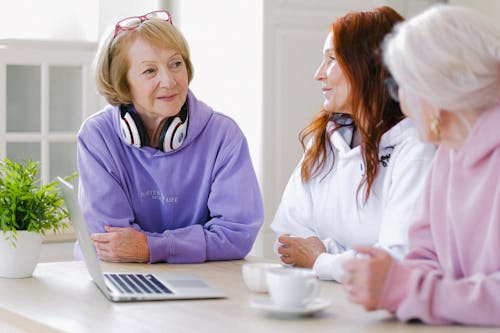 Cheerful aged women in trendy clothes sitting at table with cups of tea and laptop and communicating at home in daytime