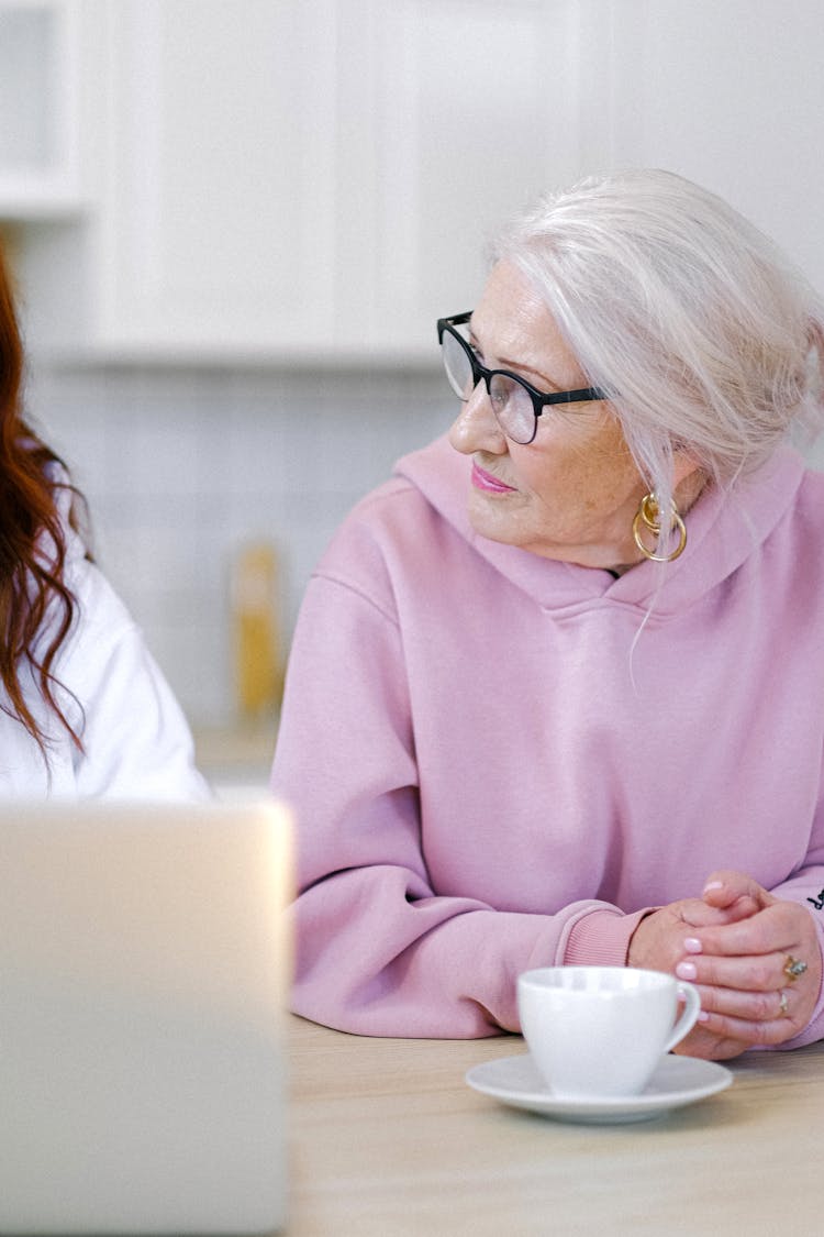 Pensive Aged Woman Sitting At Table With Friend In Light Room