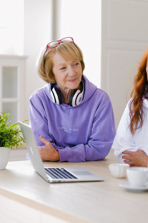 Cheerful senior woman with headphones in stylish hoodie sitting at table with opened netbook and cups of tea while talking with friends in room