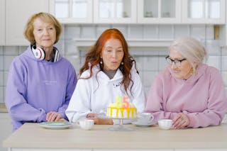 Cheerful aged woman with long red hair in white hoodie blowing out candles on birthday cake during celebration anniversary with friends at home