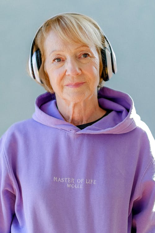 Senior female in trendy hoodie and headphones on blue background listening to music