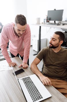 Two young men discussing work on a laptop in an office setting.