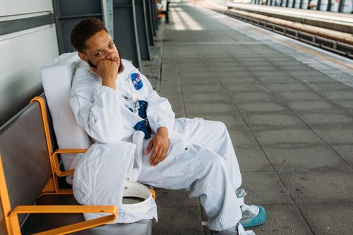 Free Man in astronaut costume sitting at an empty train station, evoking a sense of waiting and travel. Stock Photo