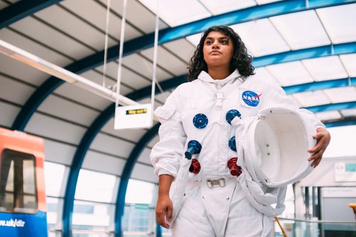 Free A woman in a space suit stands confidently in a modern subway station, blending science fiction with urban life. Stock Photo
