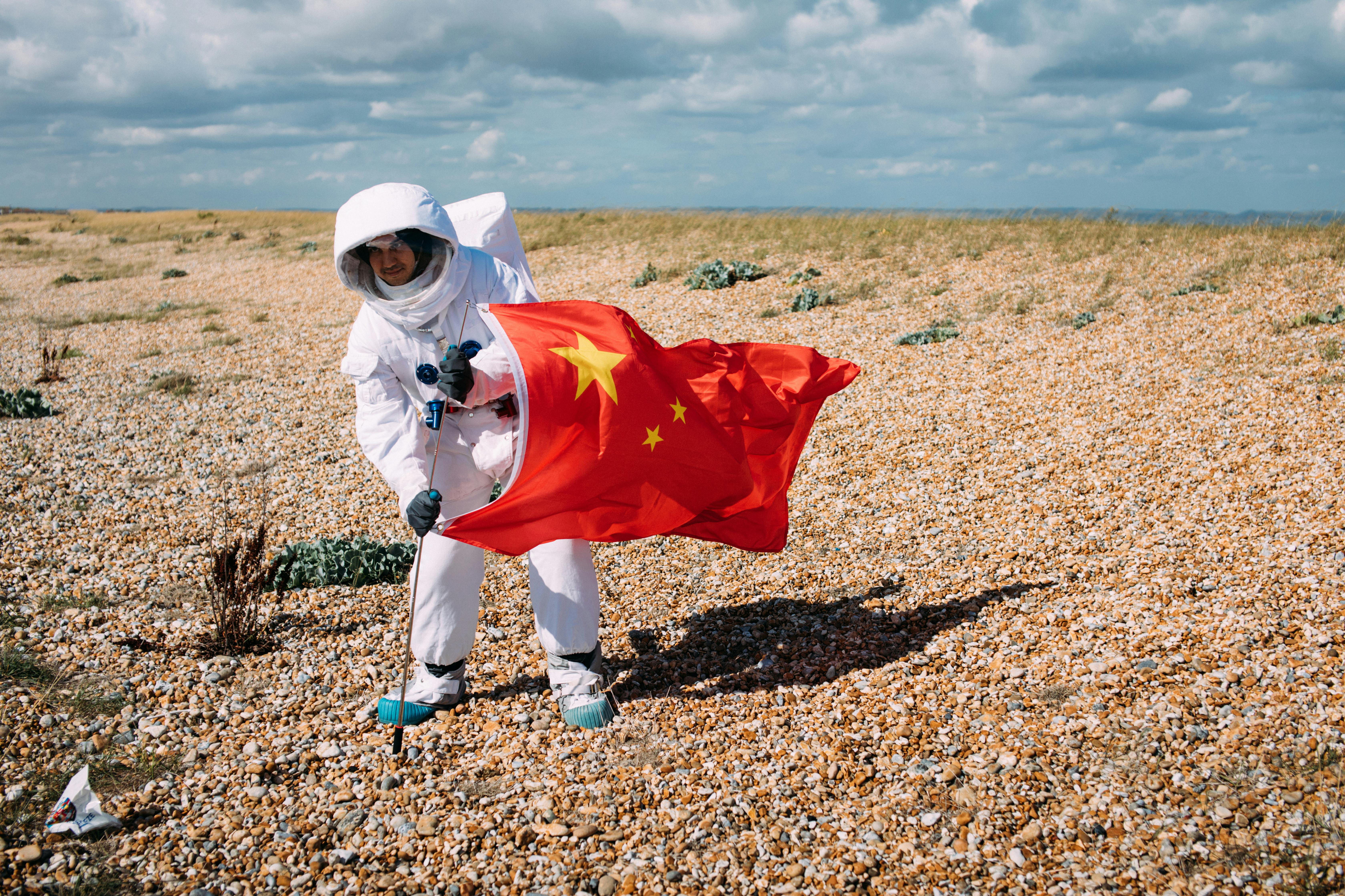 在岩石沙漠中，穿着太空服的人举着中国国旗，描绘了宇航员的场景