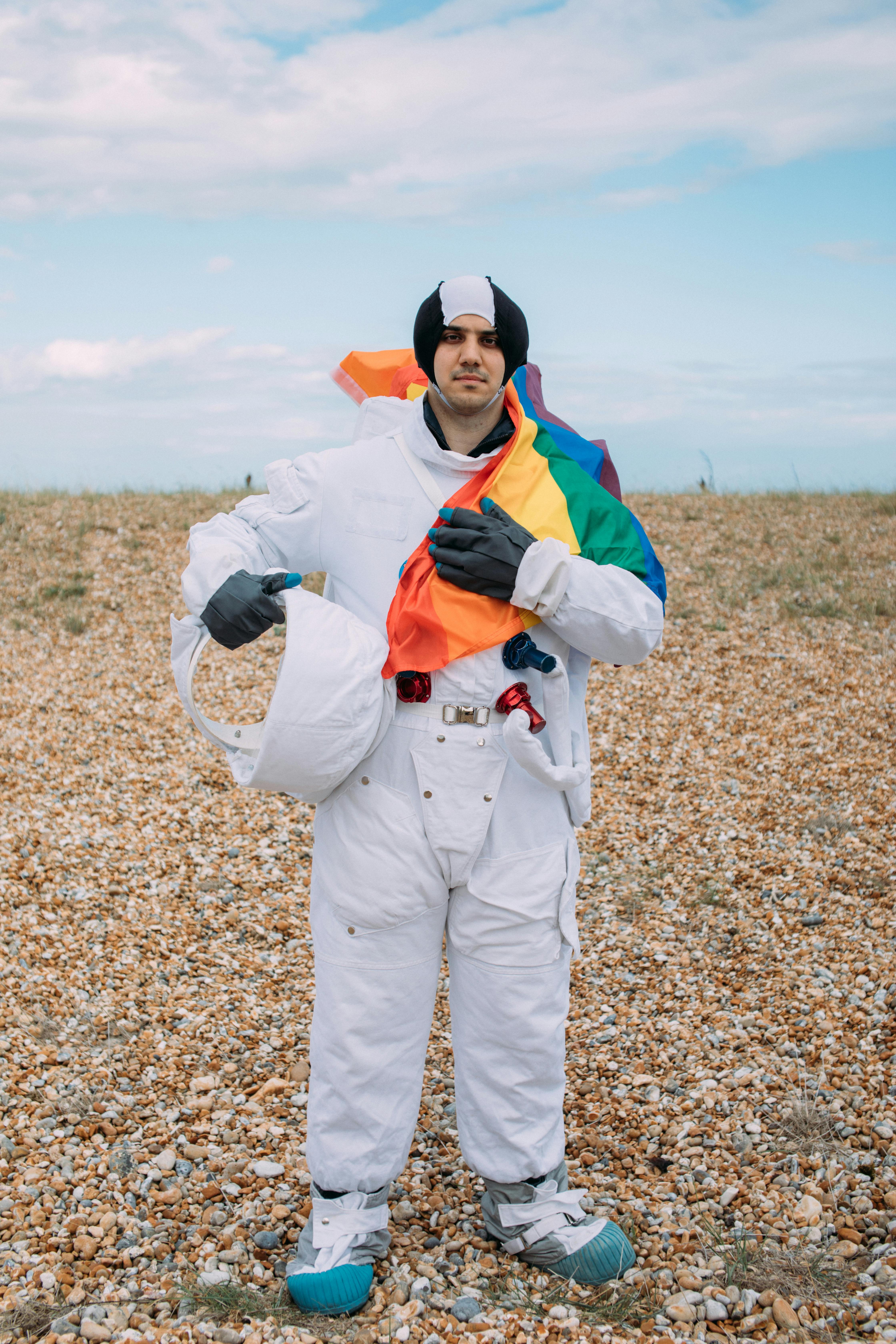 一个穿着太空服的成年人，在晴朗的天空下，举着五颜六色的旗帜站在鹅卵石景观上