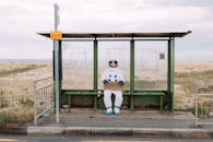 一名宇航员穿着宇航服，坐在乡村公交车站，举着一块写着“地球”的牌子