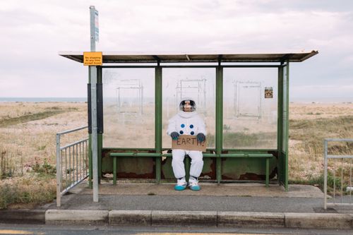 Free An astronaut in a spacesuit sits at a rural bus stop holding a sign reading 'Earth.' Stock Photo