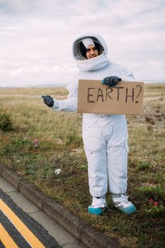一个穿着太空服的宇航员在乡村公路上搭便车，手里举着一个写着“地球？”的纸板