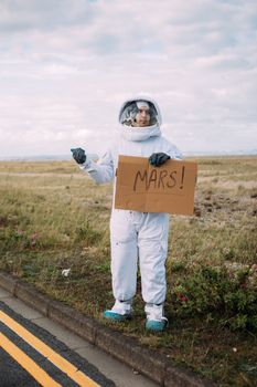 一个身穿火星标志宇航服的人，在一条荒无人烟的道路上搭便车
