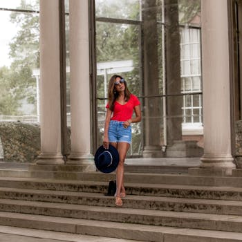 Full body of young female in sunglasses with trendy hat on concrete steps of building with tall columns