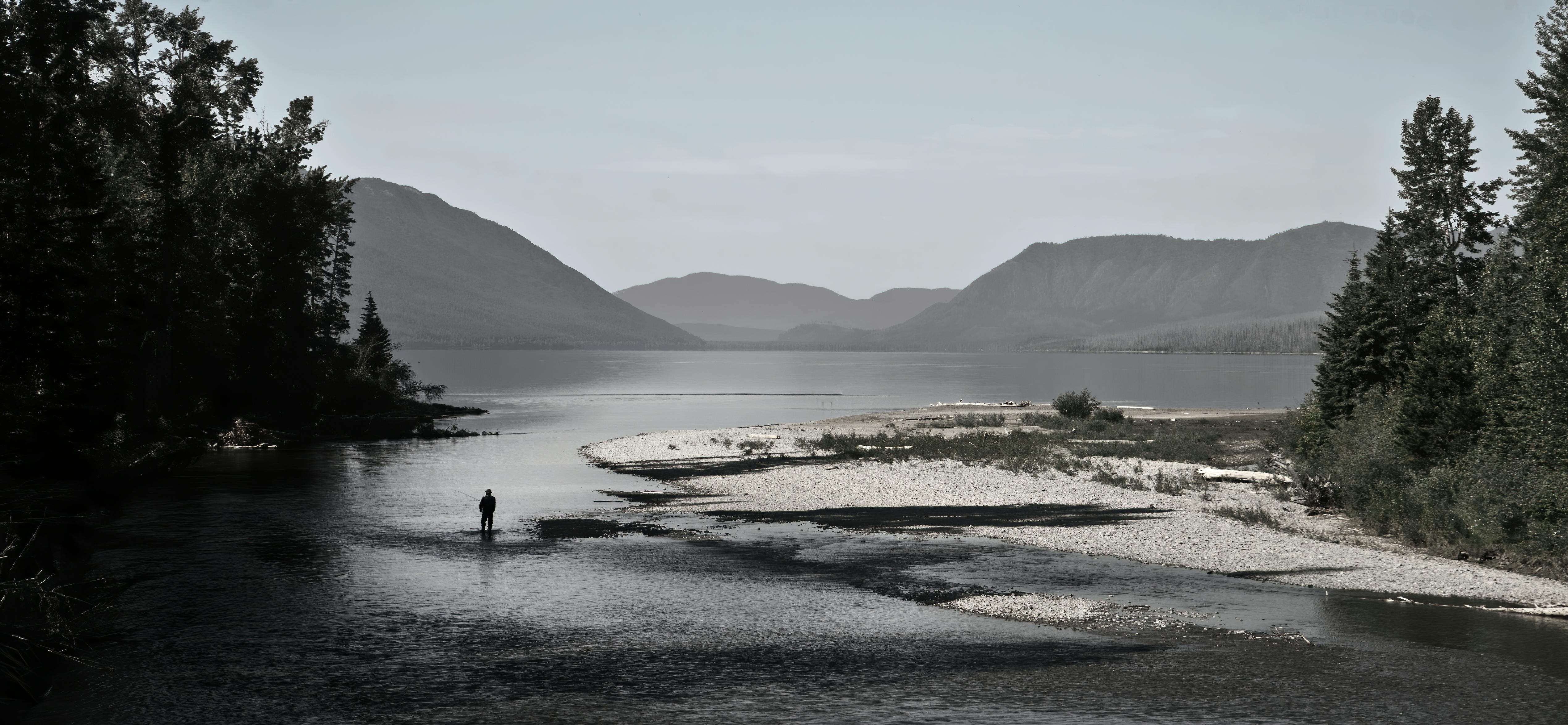 A peaceful lake view with a person fishing, surrounded by mountains and trees in Montana.