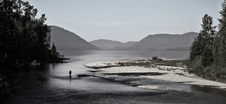A peaceful lake view with a person fishing, surrounded by mountains and trees in Montana.