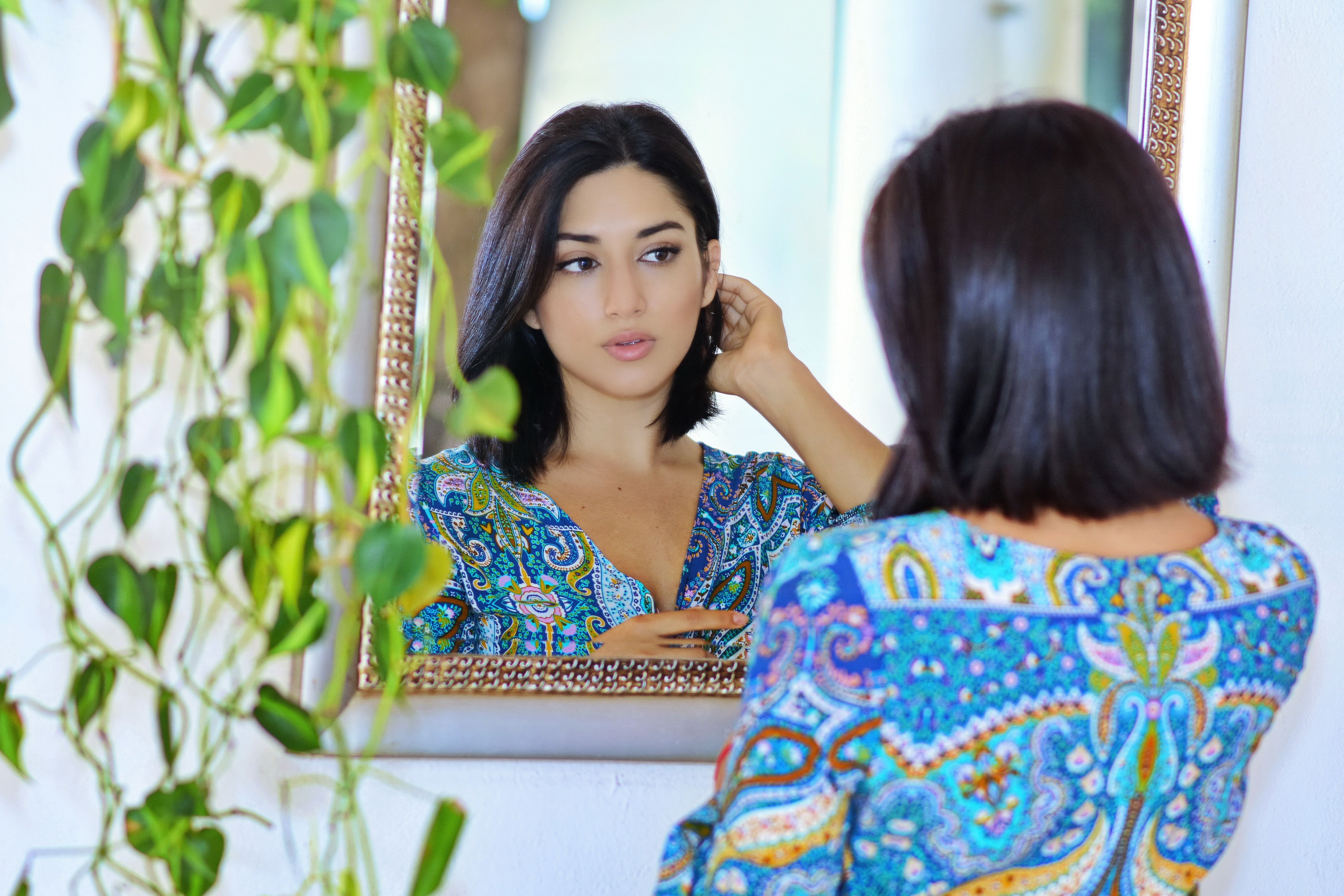 A woman adjusting her earrings, reflected in a mirror indoors with green plants.