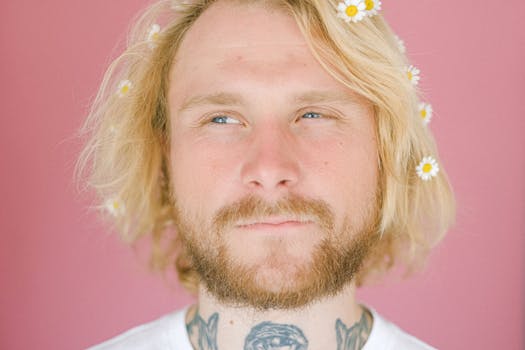 在粉色背景下，一位头发上插着雏菊的金发男子微笑着，展现出无忧无虑的轻松氛围