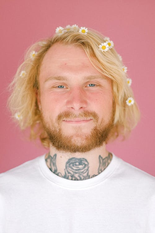 Smiling young man with flowers in blond hair looking at camera on pink background