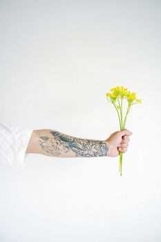 一位身着白衬衫、纹身的匿名者站在灰色背景前，身上放着一束新鲜的黄色植物