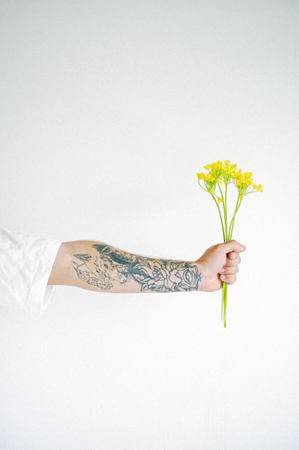 Crop anonymous person with tatoo in white shirt standing with bunch of fresh yellow plant against gray background