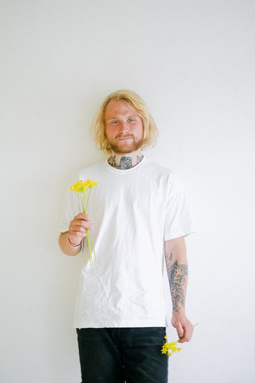 Free Sincere adult tattooed male in casual wear with blossoming flowers looking at camera in daytime Stock Photo