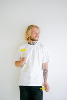 A blonde man with tattoos holding yellow flowers against a white background, looking contemplative.