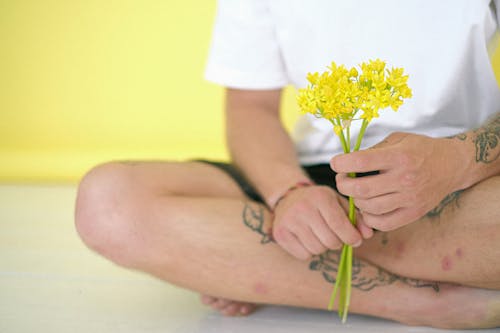 Free Crop anonymous barefoot tattooed male with blossoming flower bouquet sitting with crossed legs on yellow background Stock Photo