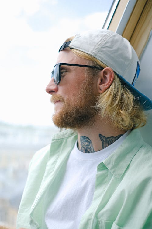 Free Adult dreamy hipster male in cap and sunglasses looking away near window in daylight Stock Photo