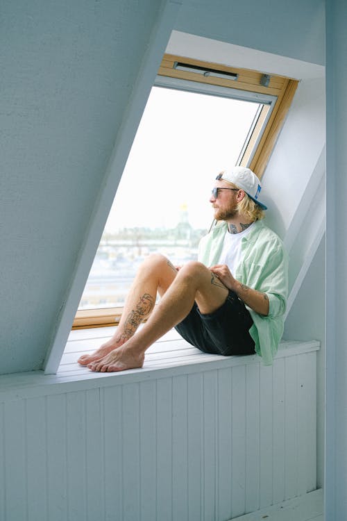 Free Dreamy barefoot hipster male in sunglasses and casual clothes looking out window in daytime Stock Photo