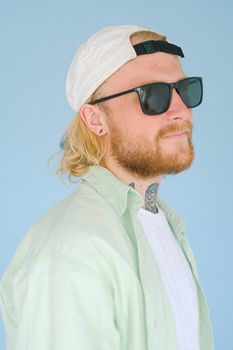 蓝色背景下，一位留着红色胡须、戴着太阳镜和帽子的时尚金发男子肖像，身上还有纹身