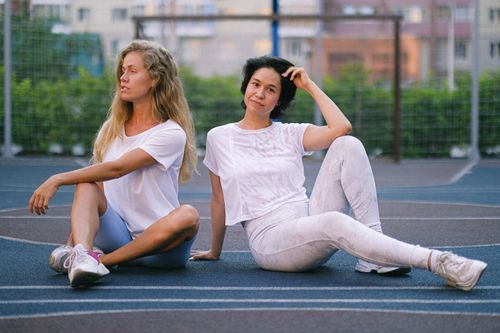 Free Young dreamy blond sportswoman sitting with crossed legs near female partner in sports clothes after working out in town Stock Photo