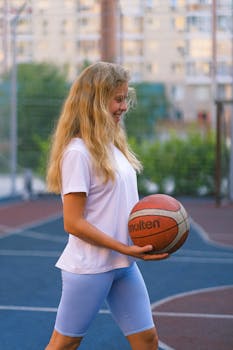 Side view of smiling young female in sportswear playing with ball on sports ground near fence in daytime