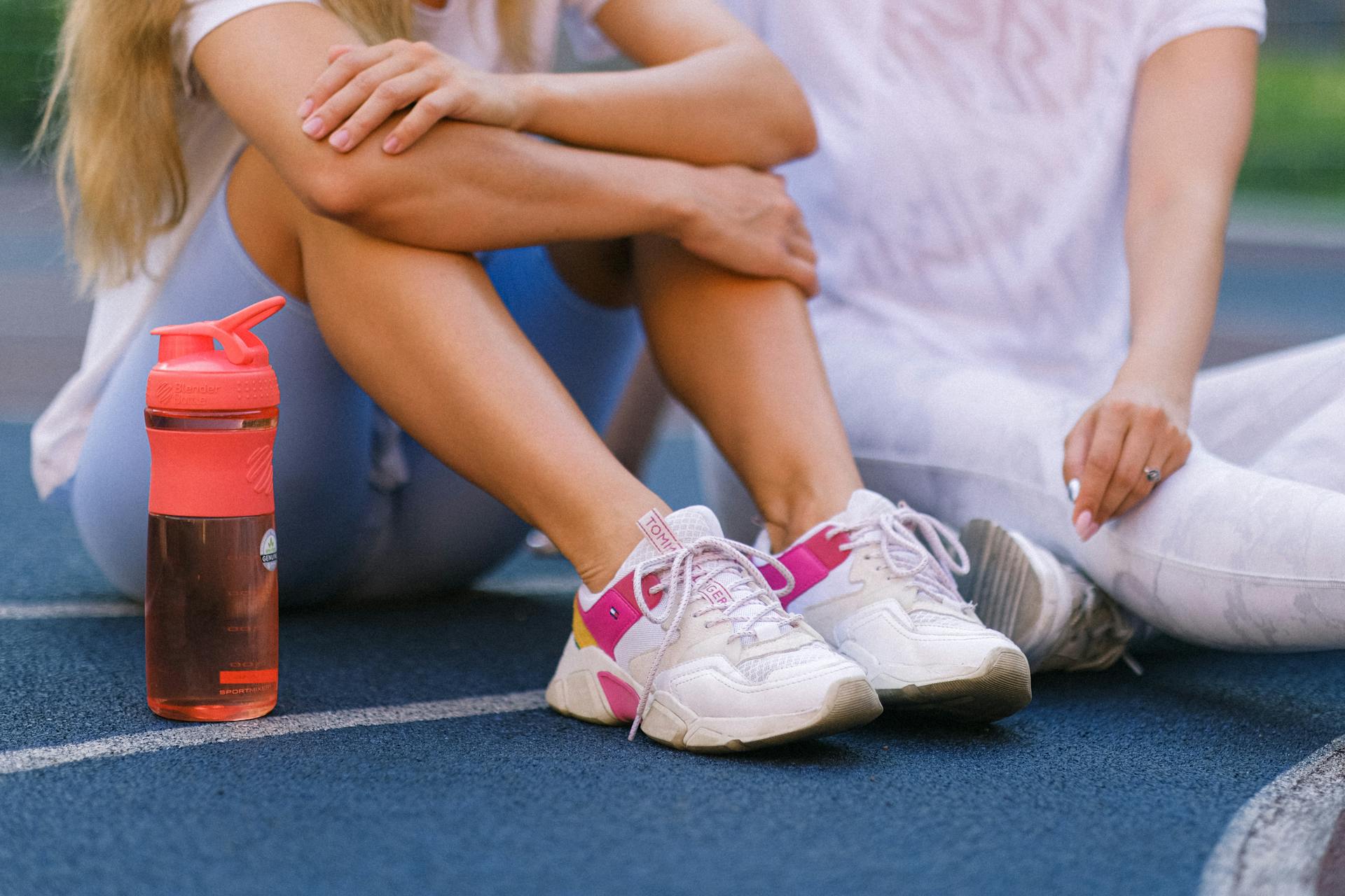 Crop anonymous female friends in activewear sitting on sports ground after training near bottle with water in daytime